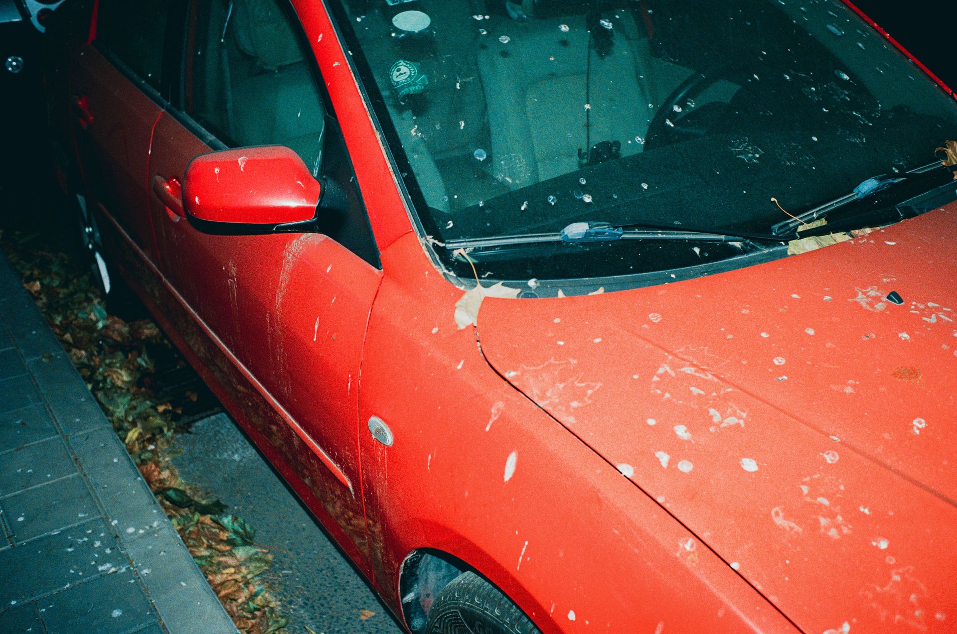 A red car with a dirty hood parked on a leaf-strewn street, suggesting neglect.